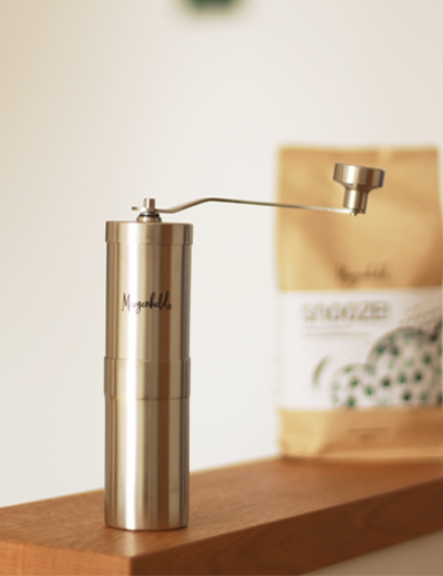 Manuelle Edelstahl-Kaffeemühle von Morgenheld mit langem Kurbelgriff, im Hintergrund eine stilvolle „SNOOZE!!“-Kaffeeverpackung – für bewusste Kaffeezubereitung mit Charakter.
