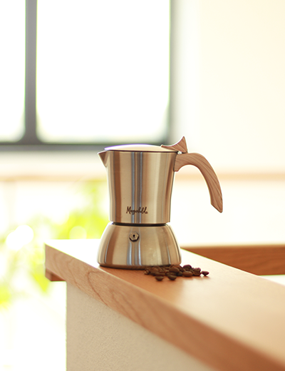 Espressokocher von Morgenheld aus Edelstahl mit Holzgriff auf einem sonnendurchfluteten Fensterbrett, umgeben von frischen Kaffeebohnen – stilvoller Genussmoment für Zuhause.