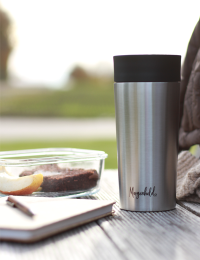 Edelstahl-Thermobecher mit Morgenheld-Logo auf Holzuntergrund im Grünen, daneben Apfelstück, Schokokuchen und Notizbuch – perfekter Begleiter für entspannte Outdoor-Momente