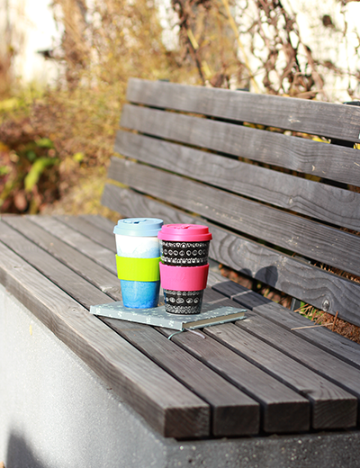 Zwei farbenfrohe Coffee to Go von Morgenheld auf einer Holzbank im Grünen – nachhaltige Kaffeepause mit hochwertigem Porzellanbecher