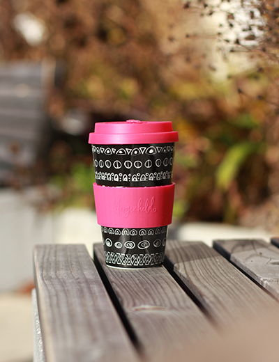 Coffee to go mit pinkem Deckel und grafischem Schwarz-Weiß-Muster auf Holzbank im Grünen – nachhaltiger Kaffeegenuss für unterwegs.